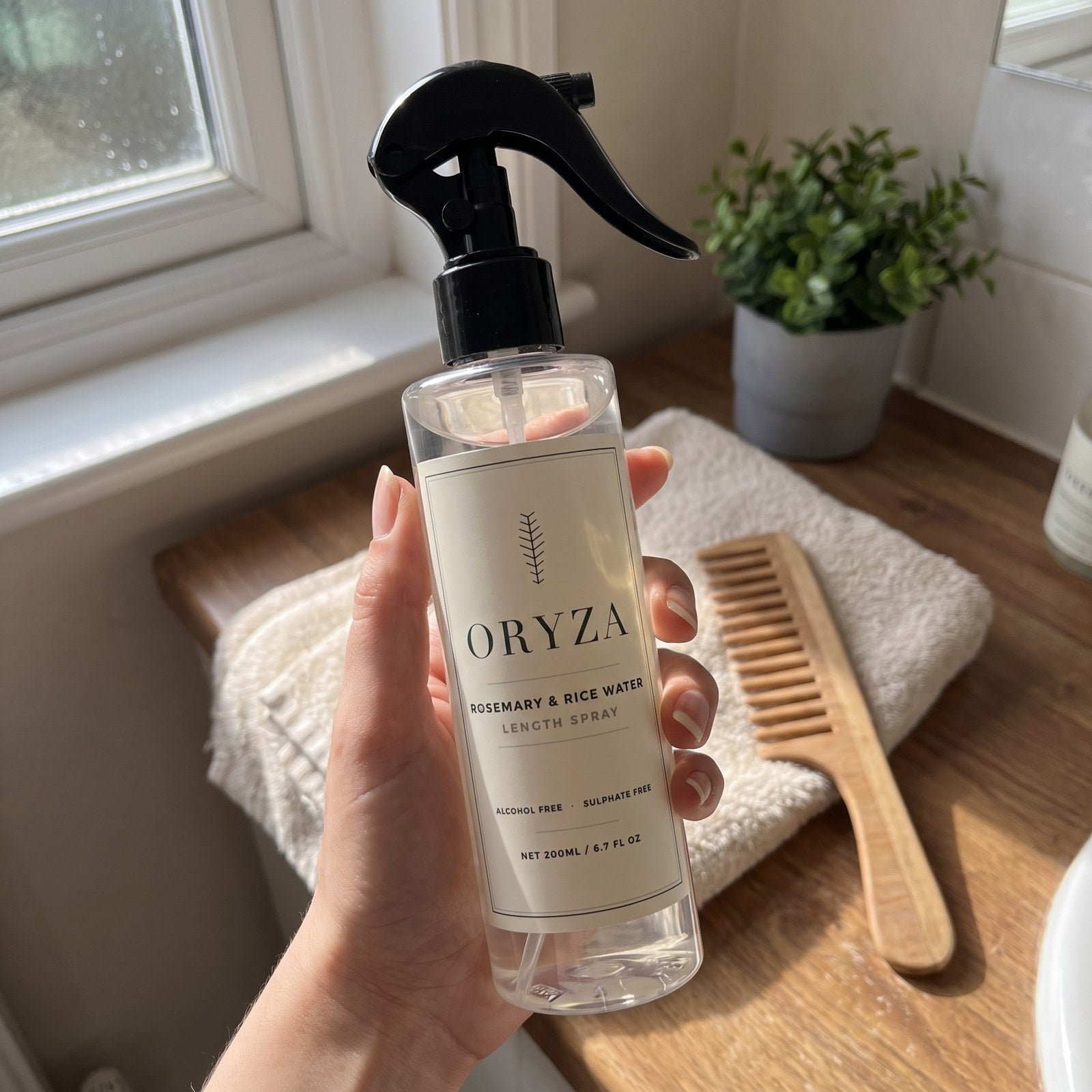 A hand holding the ORYZA bottle on a bathroom counter beside a wooden comb and folded towel